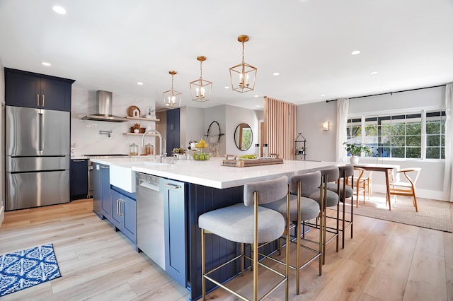 kitchen featuring decorative light fixtures, a kitchen breakfast bar, a large island with sink, stainless steel appliances, and wall chimney exhaust hood