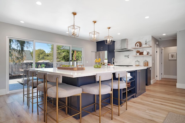 kitchen featuring blue cabinetry, wall chimney exhaust hood, a breakfast bar, decorative light fixtures, and light hardwood / wood-style flooring