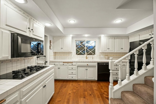 kitchen with wood finished floors, a sink, tile countertops, black appliances, and white cabinets