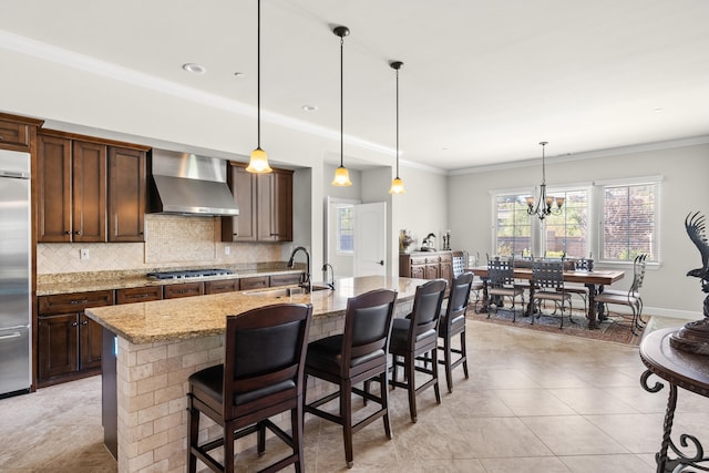 kitchen with a center island with sink, sink, wall chimney range hood, hanging light fixtures, and light stone countertops