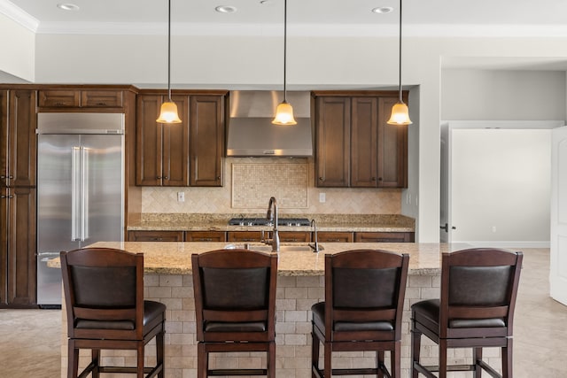 kitchen with a center island with sink, stainless steel appliances, decorative light fixtures, light stone counters, and wall chimney range hood