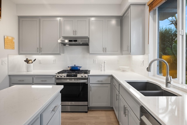 kitchen with stainless steel appliances, gray cabinets, decorative backsplash, a sink, and under cabinet range hood