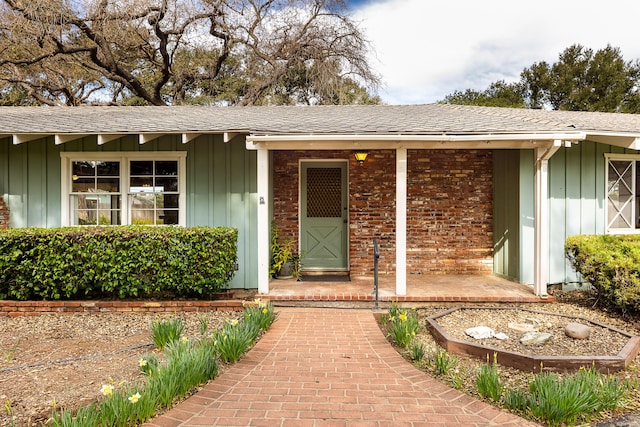 doorway to property featuring covered porch, roof with shingles, board and batten siding, and brick siding