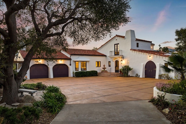 mediterranean / spanish-style home featuring stucco siding, an attached garage, a balcony, driveway, and a tiled roof