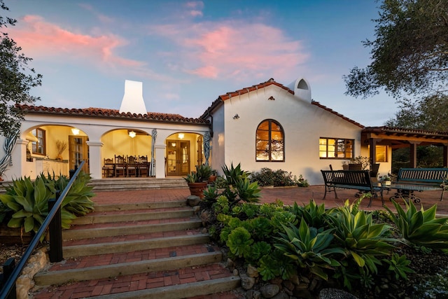 back of house at dusk featuring french doors, a tile roof, and stucco siding