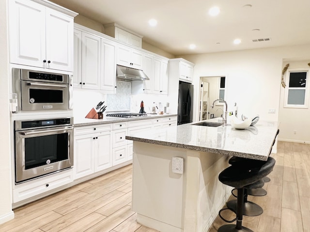 kitchen with light wood-style flooring, a kitchen island with sink, a sink, under cabinet range hood, and stainless steel appliances