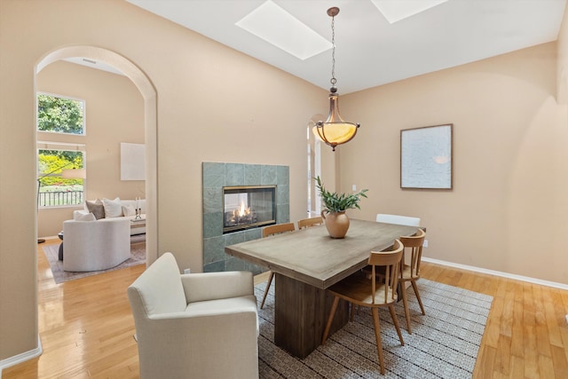 dining area featuring arched walkways, a skylight, baseboards, light wood finished floors, and a tiled fireplace