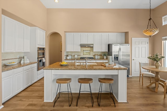 kitchen featuring arched walkways, appliances with stainless steel finishes, a kitchen island with sink, a sink, and under cabinet range hood