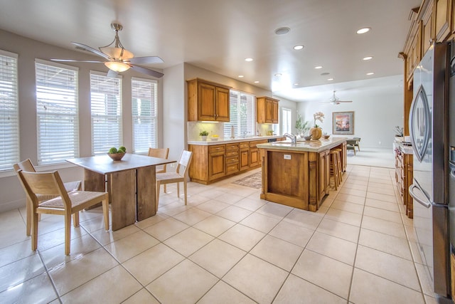 kitchen with brown cabinets, a center island with sink, light tile patterned floors, light countertops, and a ceiling fan