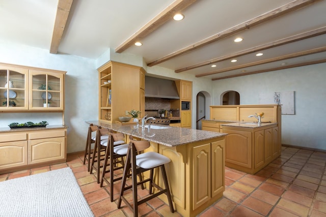 kitchen featuring arched walkways, beam ceiling, a breakfast bar area, wall chimney range hood, and black microwave