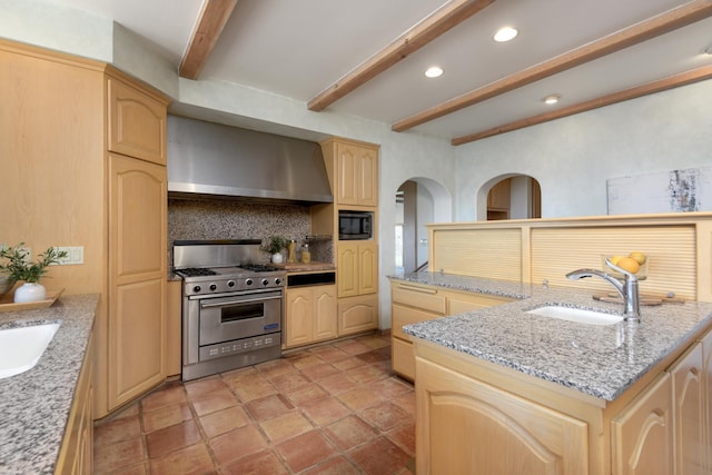 kitchen with a sink, beamed ceiling, light brown cabinetry, wall chimney exhaust hood, and stainless steel range