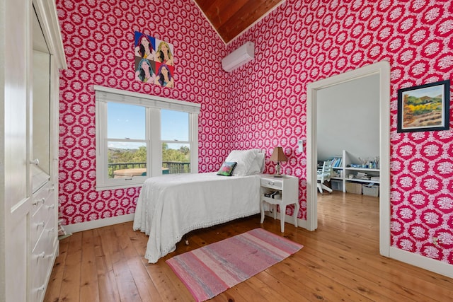 bedroom with a wall unit AC, wood-type flooring, wallpapered walls, baseboards, and vaulted ceiling