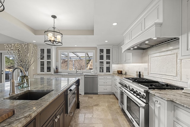 kitchen featuring stone tile floors, a sink, appliances with stainless steel finishes, a tray ceiling, and glass insert cabinets