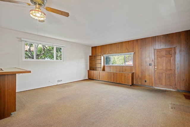 unfurnished living room with a wealth of natural light, wood walls, carpet, and ceiling fan