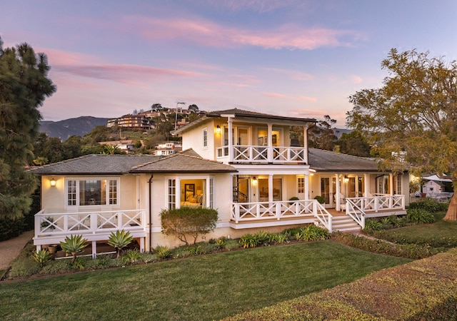 back of property at dusk featuring stucco siding, a balcony, a porch, and a lawn