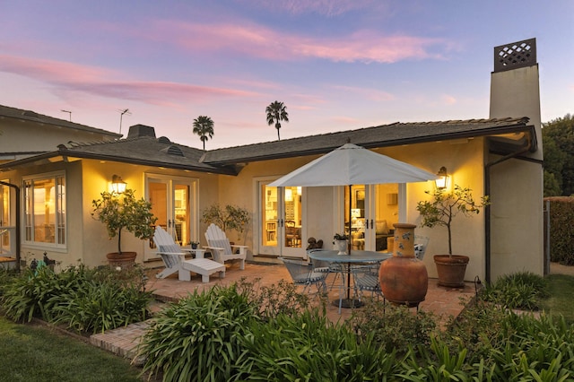 back of house at dusk with a patio area, stucco siding, french doors, and a chimney