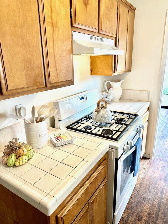 kitchen with tile counters, under cabinet range hood, white gas range, wood finished floors, and brown cabinetry