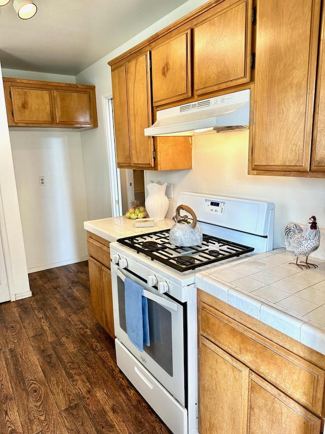 kitchen with brown cabinets, under cabinet range hood, dark wood finished floors, white gas range oven, and tile countertops