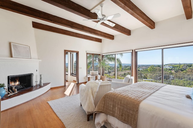 bedroom with ceiling fan, baseboards, a lit fireplace, beam ceiling, and wood-type flooring