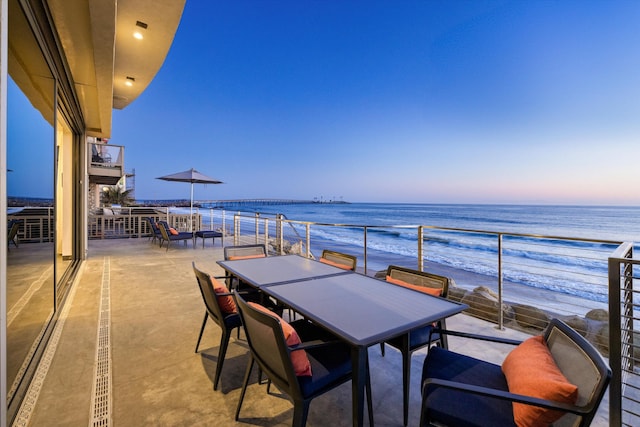 balcony at dusk featuring a beach view, a water view, and outdoor dining area