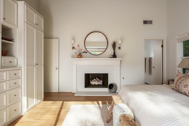 bedroom with a fireplace with raised hearth, light wood finished floors, a towering ceiling, and visible vents