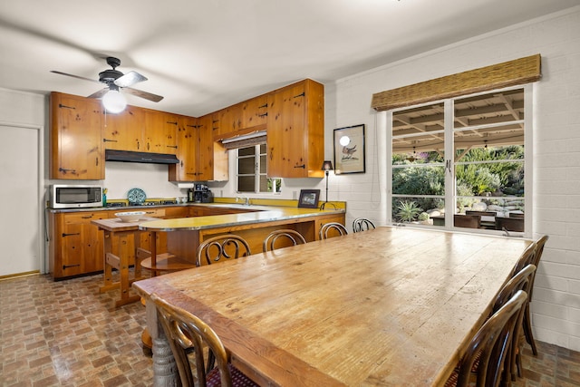 kitchen with brick floor, brown cabinetry, ceiling fan, appliances with stainless steel finishes, and under cabinet range hood