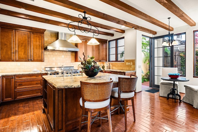 kitchen featuring a sink, an island with sink, wall chimney range hood, light stone countertops, and dark wood-type flooring