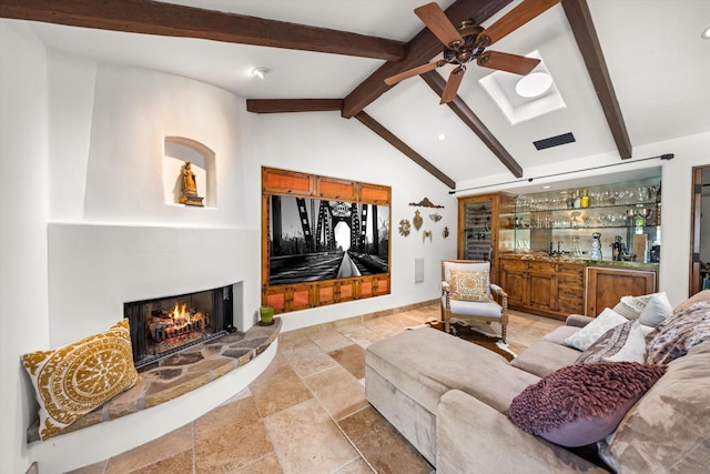 living area with a warm lit fireplace, ceiling fan, a dry bar, vaulted ceiling with beams, and stone tile flooring