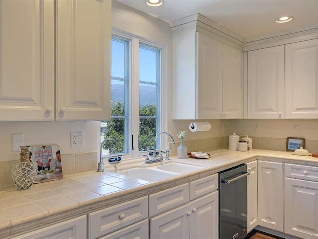 kitchen featuring stainless steel dishwasher, tile counters, a sink, white cabinetry, and recessed lighting
