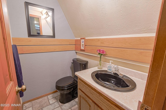 bathroom featuring stone tile flooring, a textured wall, toilet, vanity, and baseboards