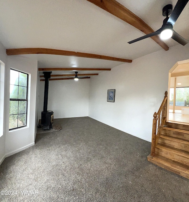 unfurnished living room featuring a wood stove, ceiling fan, stairway, and beamed ceiling