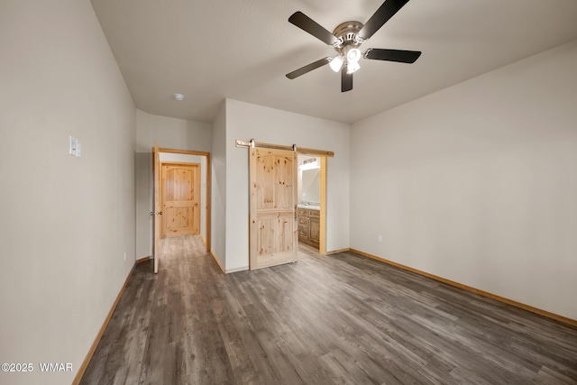 unfurnished bedroom featuring dark wood-style floors, a barn door, connected bathroom, ceiling fan, and baseboards