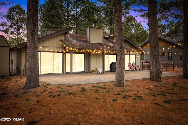 back of house at dusk with a chimney, board and batten siding, a patio area, a garage, and driveway