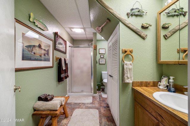 full bathroom featuring a drop ceiling, toilet, stone finish flooring, vanity, and a shower stall