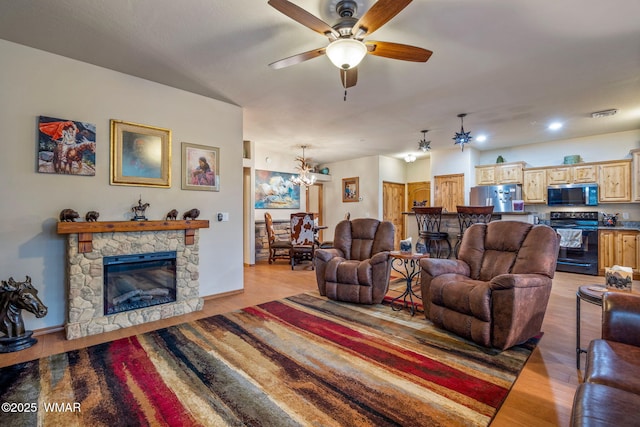 living area featuring light wood-style floors, a stone fireplace, baseboards, and ceiling fan with notable chandelier