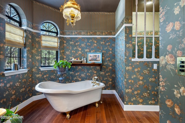 full bathroom with wood finished floors, a chandelier, a freestanding tub, baseboards, and wallpapered walls