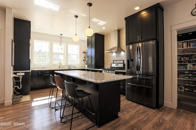 kitchen with stainless steel appliances, a kitchen island, wall chimney range hood, light stone countertops, and dark wood finished floors