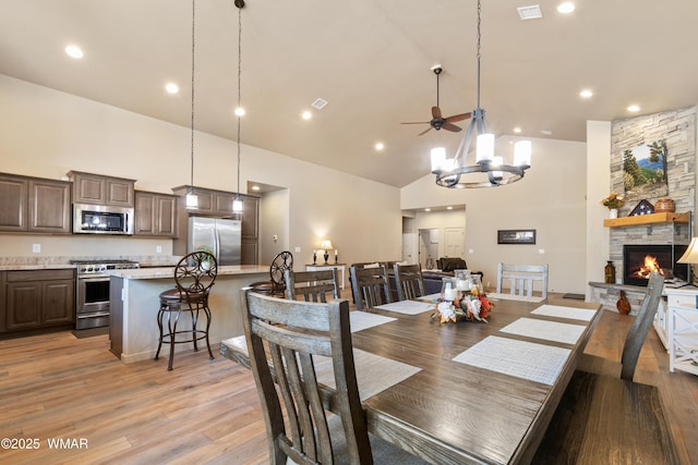 dining room featuring visible vents, a stone fireplace, light wood-style floors, high vaulted ceiling, and ceiling fan with notable chandelier