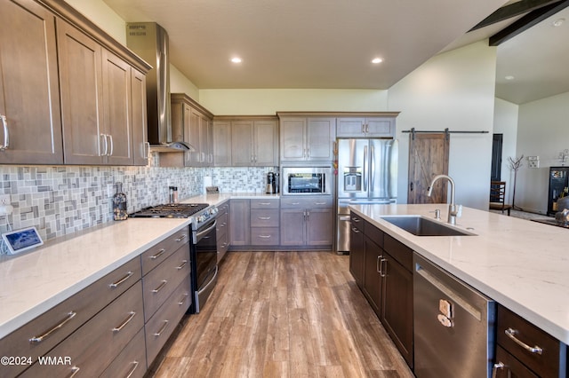 kitchen featuring a barn door, light stone counters, stainless steel appliances, a sink, and wall chimney exhaust hood