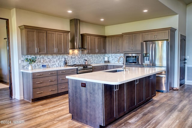 kitchen featuring stainless steel appliances, a sink, light countertops, wall chimney exhaust hood, and an island with sink