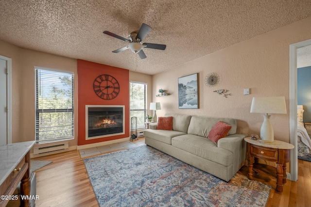 living area featuring a textured ceiling, a textured wall, baseboard heating, light wood-type flooring, and a glass covered fireplace