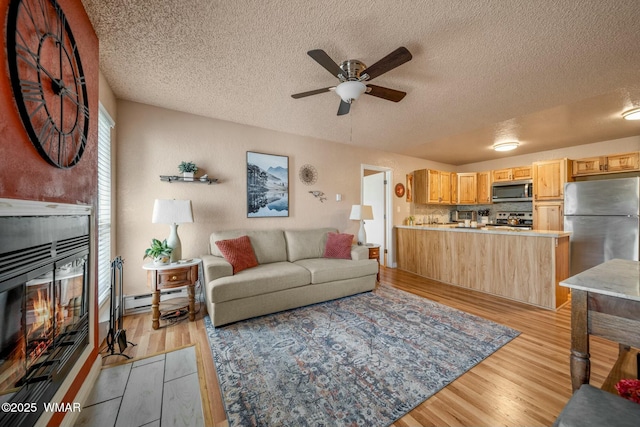 living room with light wood-style flooring, baseboard heating, a tiled fireplace, ceiling fan, and a textured ceiling