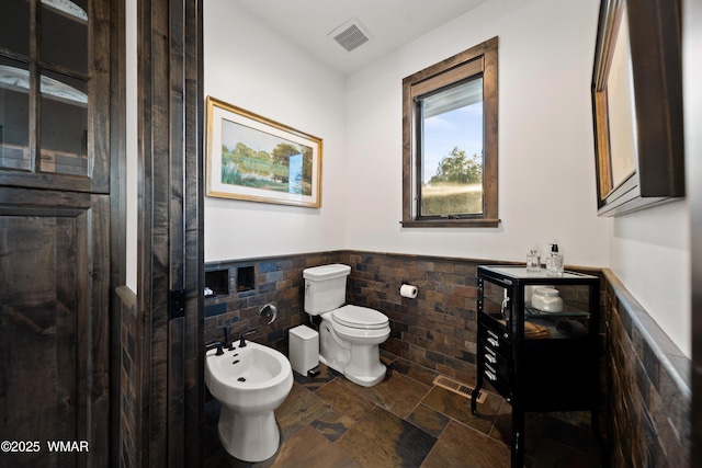 bathroom featuring tile walls, stone tile floors, toilet, wainscoting, and a bidet