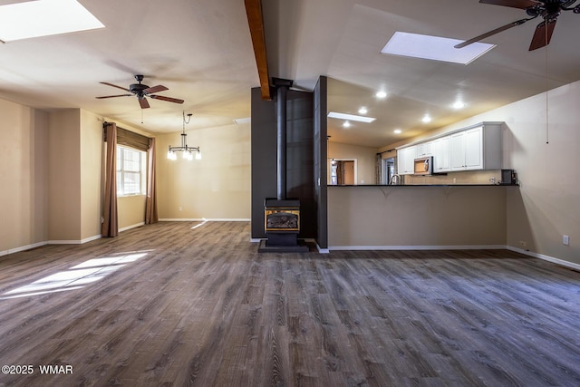 unfurnished living room with ceiling fan, vaulted ceiling with skylight, dark wood-style flooring, baseboards, and a wood stove