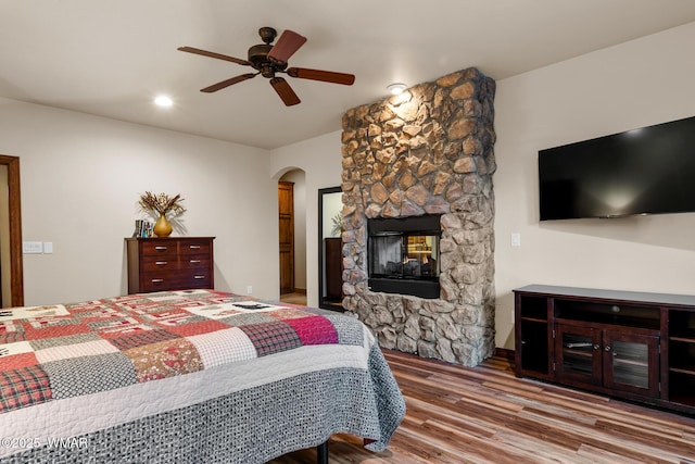 bedroom featuring a ceiling fan, arched walkways, wood finished floors, and a stone fireplace