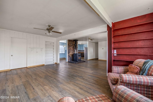 unfurnished living room featuring a wood stove, a ceiling fan, dark wood-type flooring, and beamed ceiling