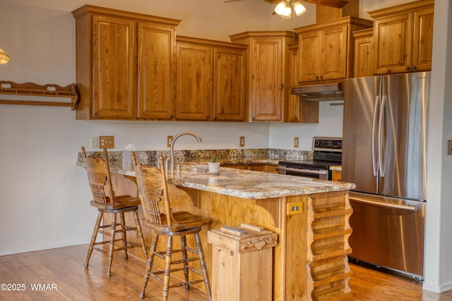 kitchen featuring brown cabinets, a peninsula, light stone countertops, stainless steel appliances, and under cabinet range hood