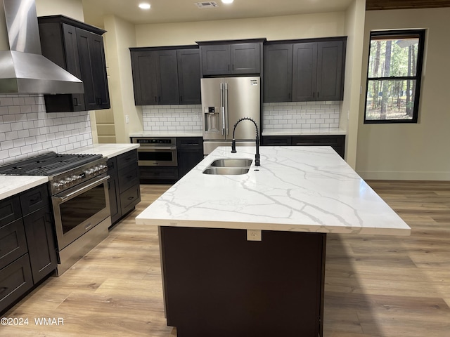 kitchen featuring premium appliances, an island with sink, wall chimney exhaust hood, light wood-type flooring, and a sink