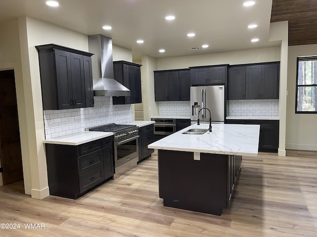 kitchen featuring dark cabinets, wall chimney exhaust hood, an island with sink, and stainless steel appliances