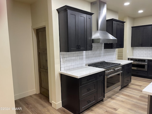 kitchen with light wood finished floors, wall chimney range hood, appliances with stainless steel finishes, and dark cabinetry